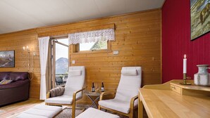 Une pièce chaleureuse aux murs en bois, avec un mur d’accent rouge, une grande fenêtre, un canapé, deux fauteuils inclinables, une petite table avec des bouteilles et des verres, et une bougie posée sur un plateau.