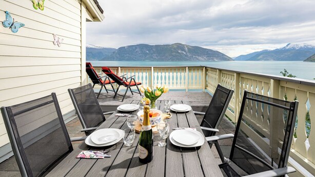 Une terrasse en bois avec une table dressée pour le repas, donnant sur un lac et offrant une vue sur des montagnes en arrière-plan.
