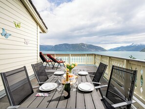 A wooden deck with a table set for dining, overlooking a lake with mountains in the background.