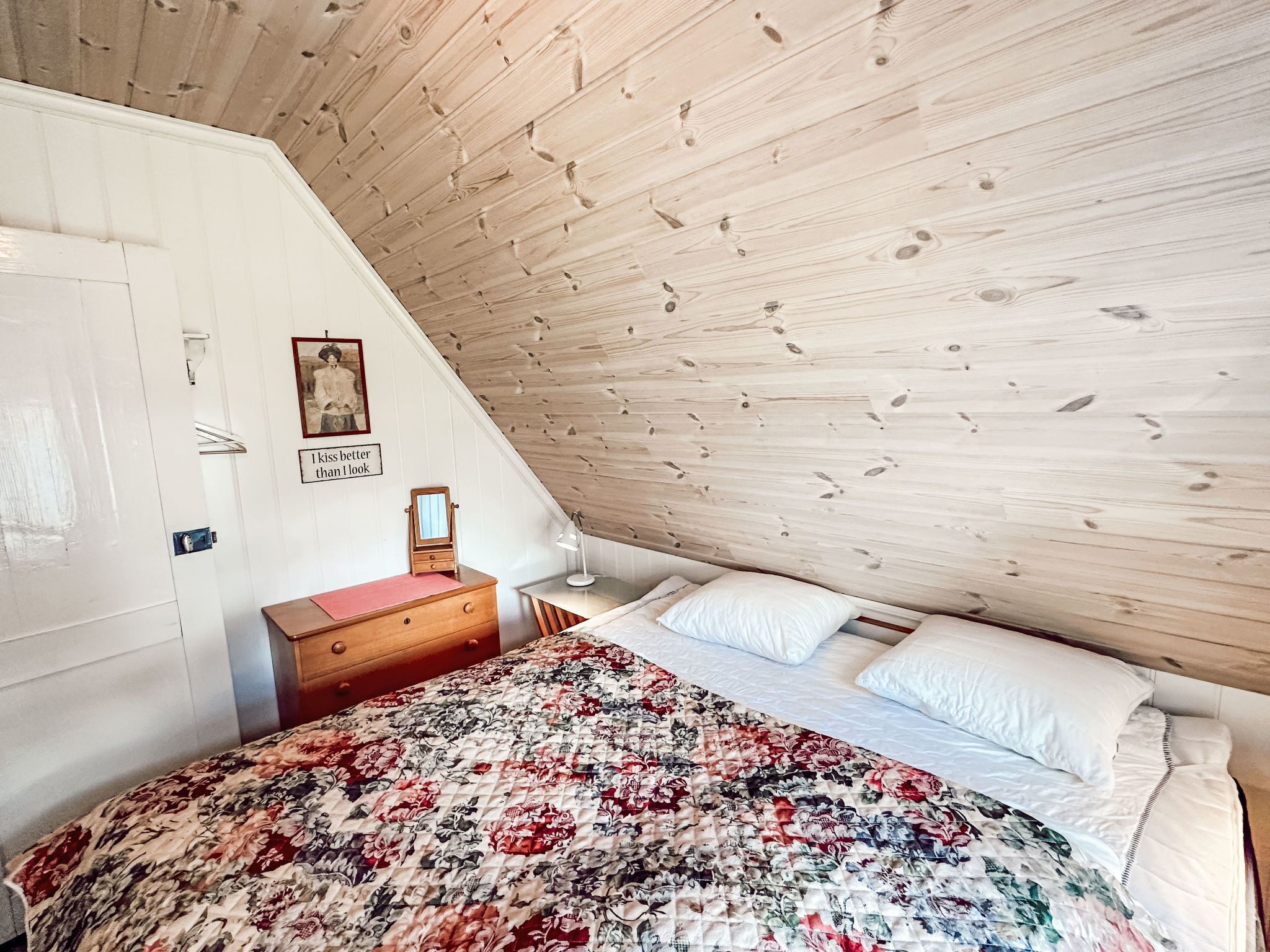 A bedroom with a sloped ceiling, a wooden dresser, a bed with a floral quilt, and a wall with a framed picture and a quote.