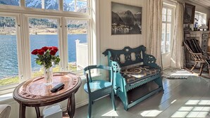 Ein Zimmer mit großem Fenster mit Blick auf einen See und Berge, ein Holztisch mit einer Vase roter Rosen, eine blaue Bank und ein gerahmtes Bild an der Wand.