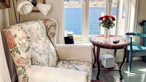 Ein gemütliches Zimmer mit einem floralen Sessel, einem runden Holztisch mit einer Vase voller roter Blumen, einem gerahmten Bild an der Wand und Seeblick durch große Fenster.