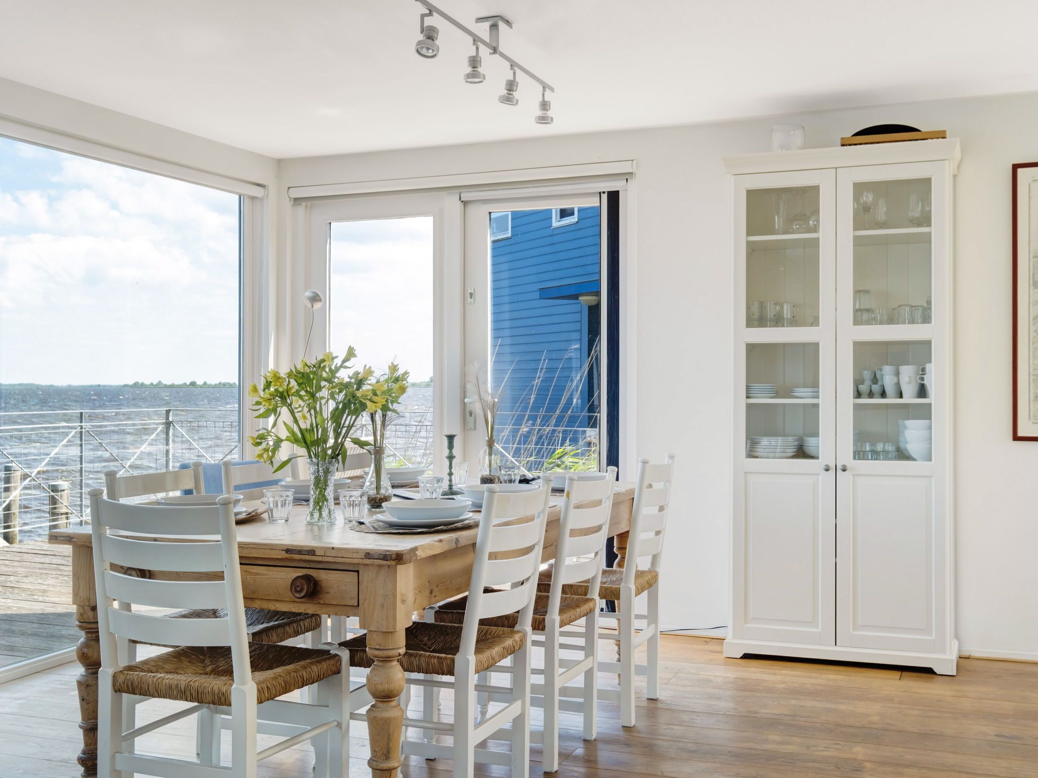 Uma sala de jantar com mesa de madeira, cadeiras brancas e vista para o mar por grandes janelas.