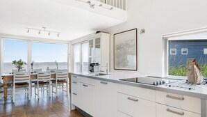 A modern kitchen with white cabinetry, a wooden dining table with chairs, and large windows offering a view of the sea.
