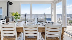 A dining area with a wooden table, white chairs, and a view of the sea through large windows.