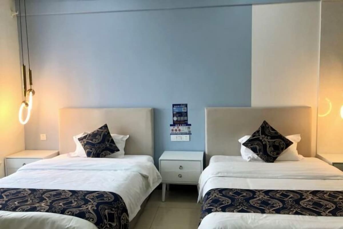 Quarto de hotel com duas camas, cada uma com colcha estampada, uma pequena mesa de cabeceira e luminária fixa na parede.