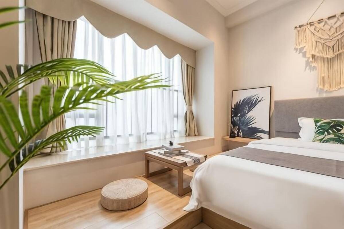 Ein Hotelzimmer mit Bett, Nachttisch, einem kleinen runden Hocker, einem Fenster mit Vorhängen und einer wandhängenden Makramee-Dekoration.