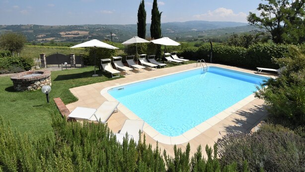 A rectangular swimming pool with a clear blue water, surrounded by a paved deck. There are several white lounge chairs and umbrellas arranged around the pool. The pool is set in a landscaped garden with neatly trimmed hedges and a variety of shrubs. In the background, there is a view of rolling hills and a clear sky. - La Corte Bricca (Trilo B) by Interhome (Oltrepò Pavese)