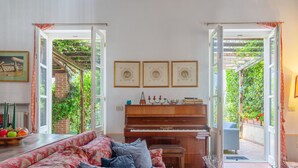 A living room with a piano, a sofa, and a dining table. There are framed pictures on the wall and a window with a view of greenery. - Ca' del Vento by Interhome (Oltrepò Pavese)