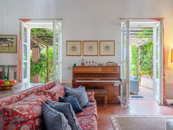 A living room with a piano, a sofa, and a dining table. There are framed pictures on the wall and a window with a view of greenery. - Ca' del Vento by Interhome (Oltrepò Pavese)