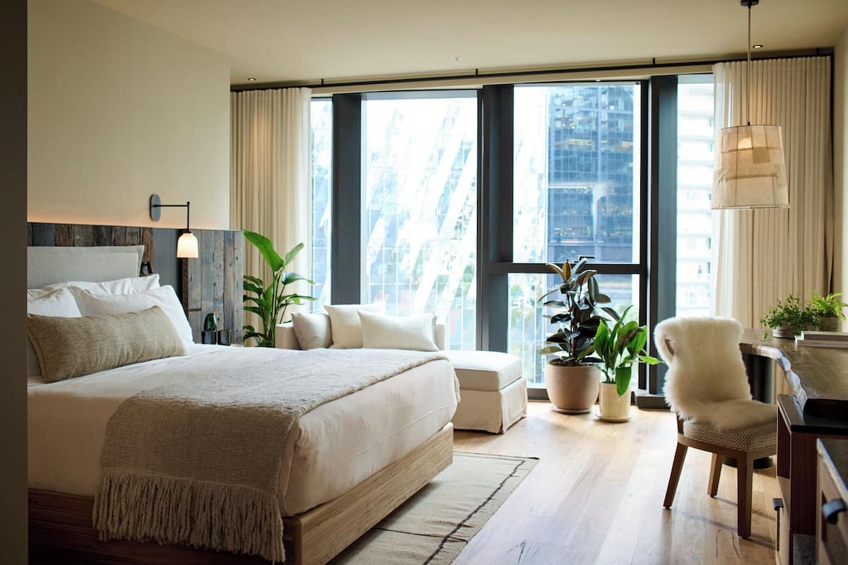 Un dormitorio moderno con una cama grande, un escritorio y una silla. Hay plantas en macetas y se ve una vista de edificios a través de la ventana.
