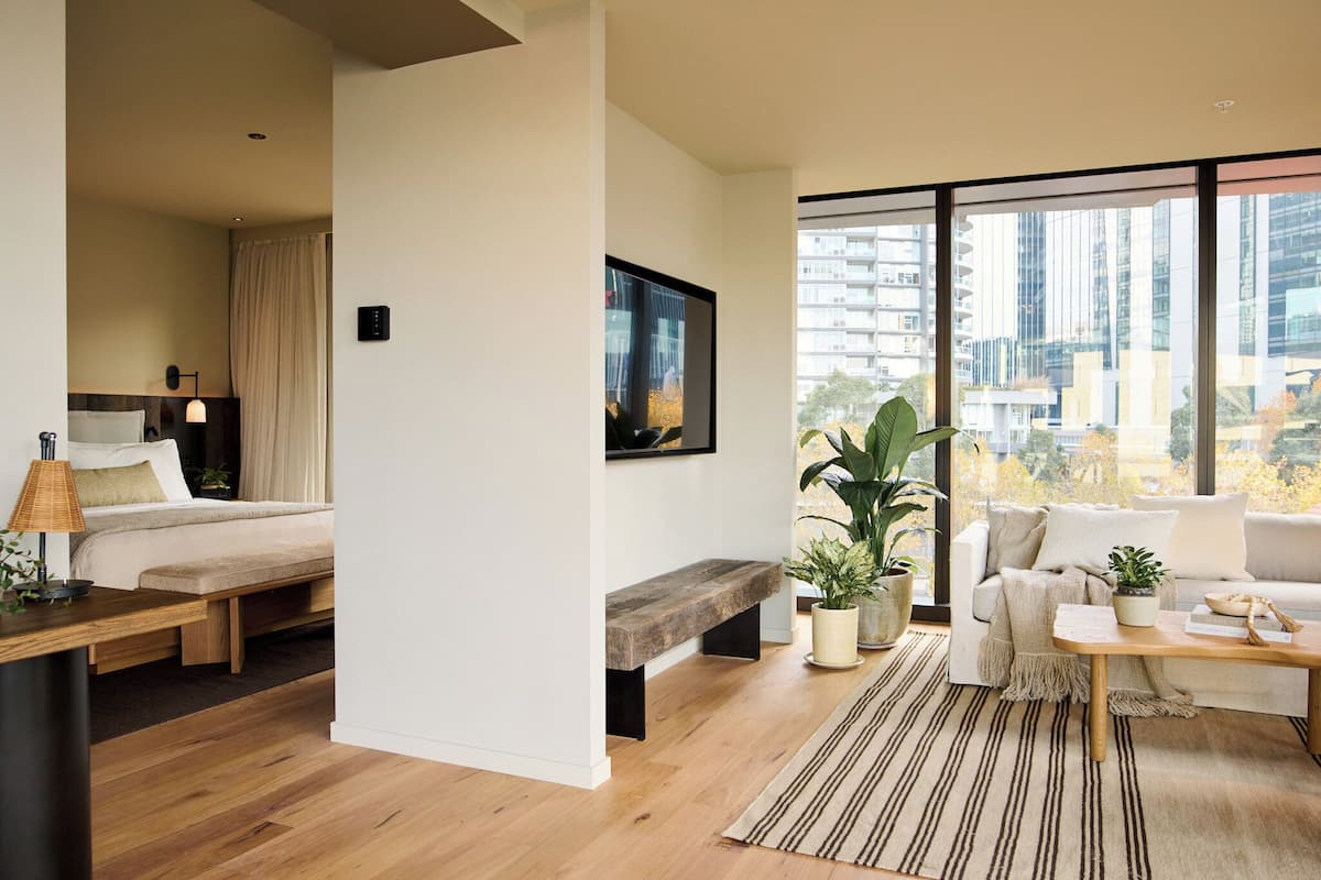 Habitación de hotel moderna con cama, sofá, mesa de centro, banco y ventanas amplias con vista a la ciudad.