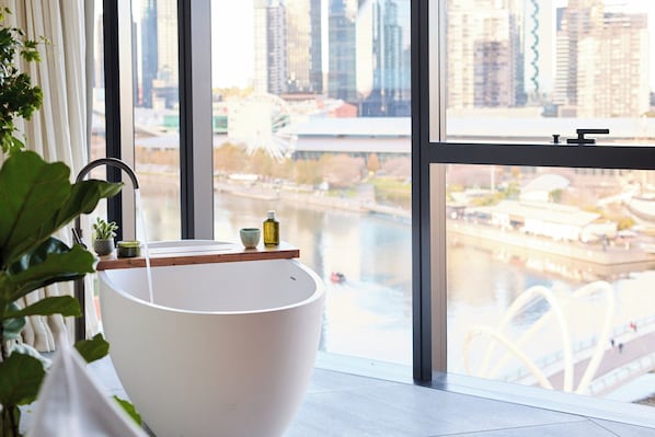 A modern bathroom with a large bathtub, a window with a city view, and a potted plant.