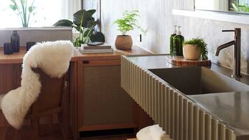 A modern kitchen with a wooden countertop, a built-in sink, and a large mirror above it. There are plants on the counter and a fluffy chair in the corner.