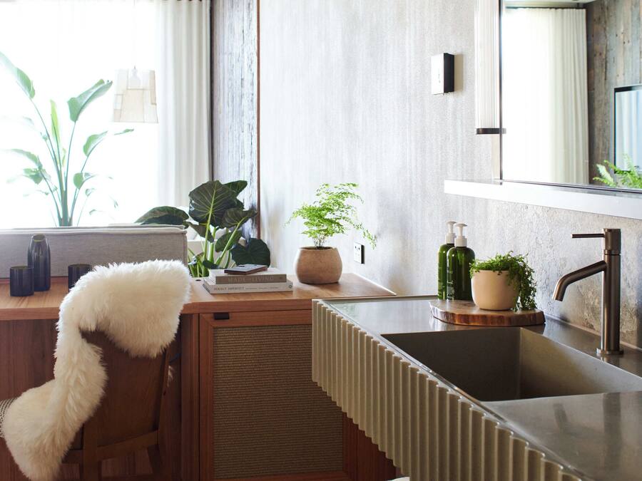 A modern kitchen with a wooden countertop, a built-in sink, and a large mirror above it. There are plants on the counter and a fluffy chair in the corner.