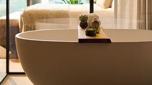 Een moderne badkamer met een groot bad, een klein tafeltje met toiletartikelen, en een uitzicht op een stadslandschap door grote ramen.