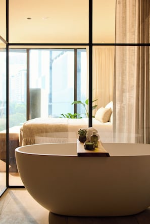 A modern bathroom with a large bathtub, a small table with toiletries, and a view of a cityscape through large windows.