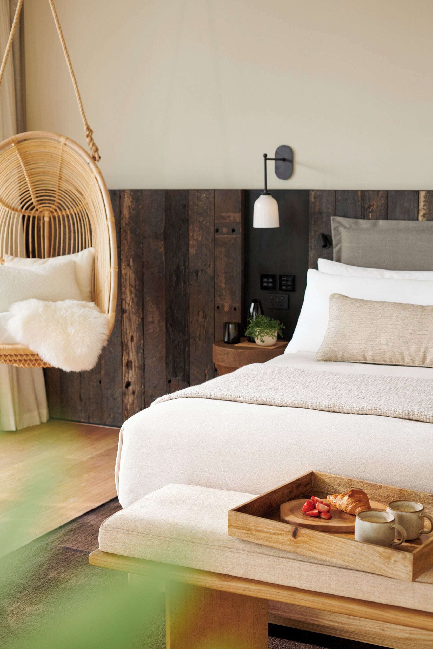 Un dormitorio con cama, mesita de noche, silla colgante, lámpara de noche y una bandeja con comida y bebidas.