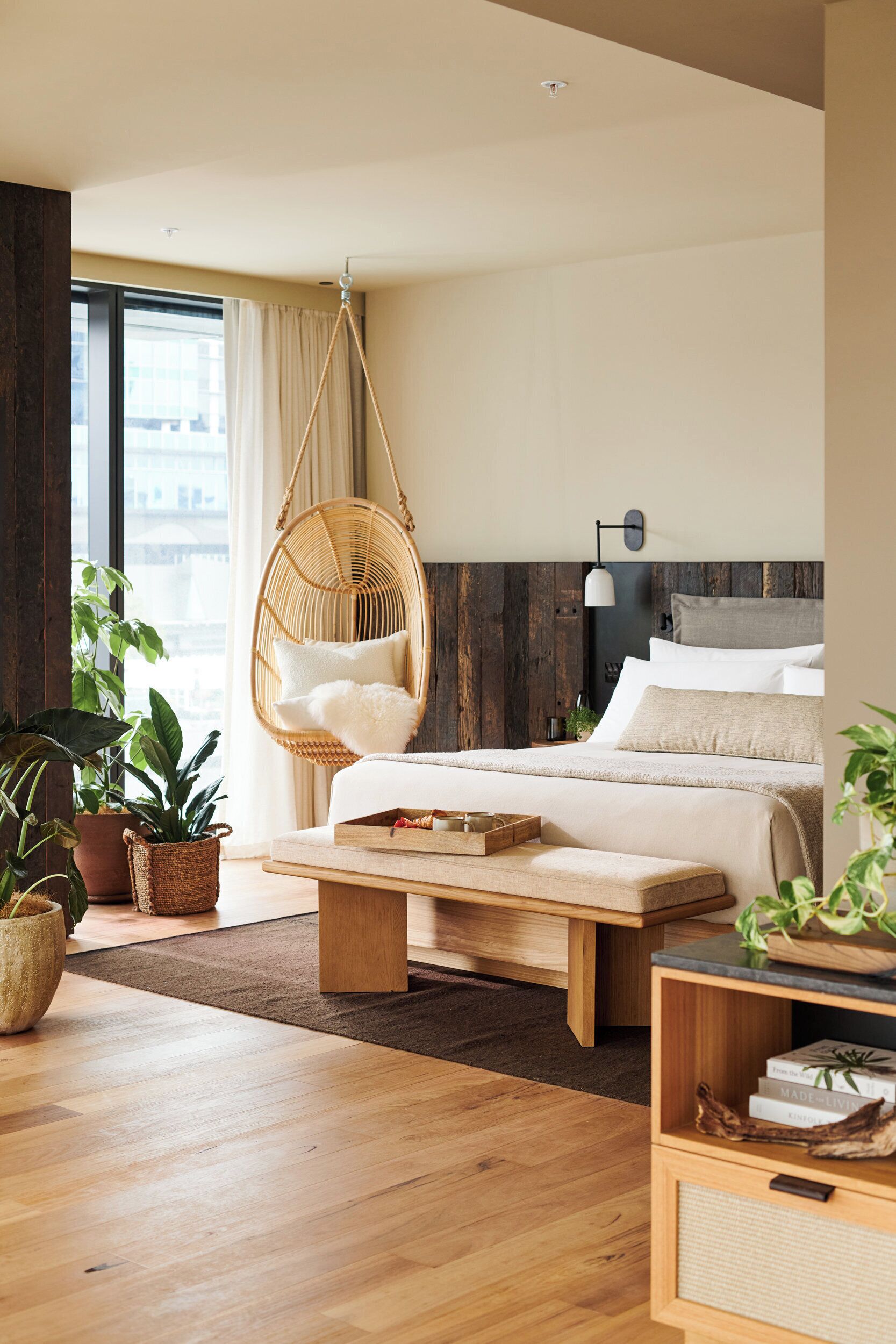 A modern bedroom with a hanging chair, a bed with white bedding, a wooden coffee table, and potted plants.
