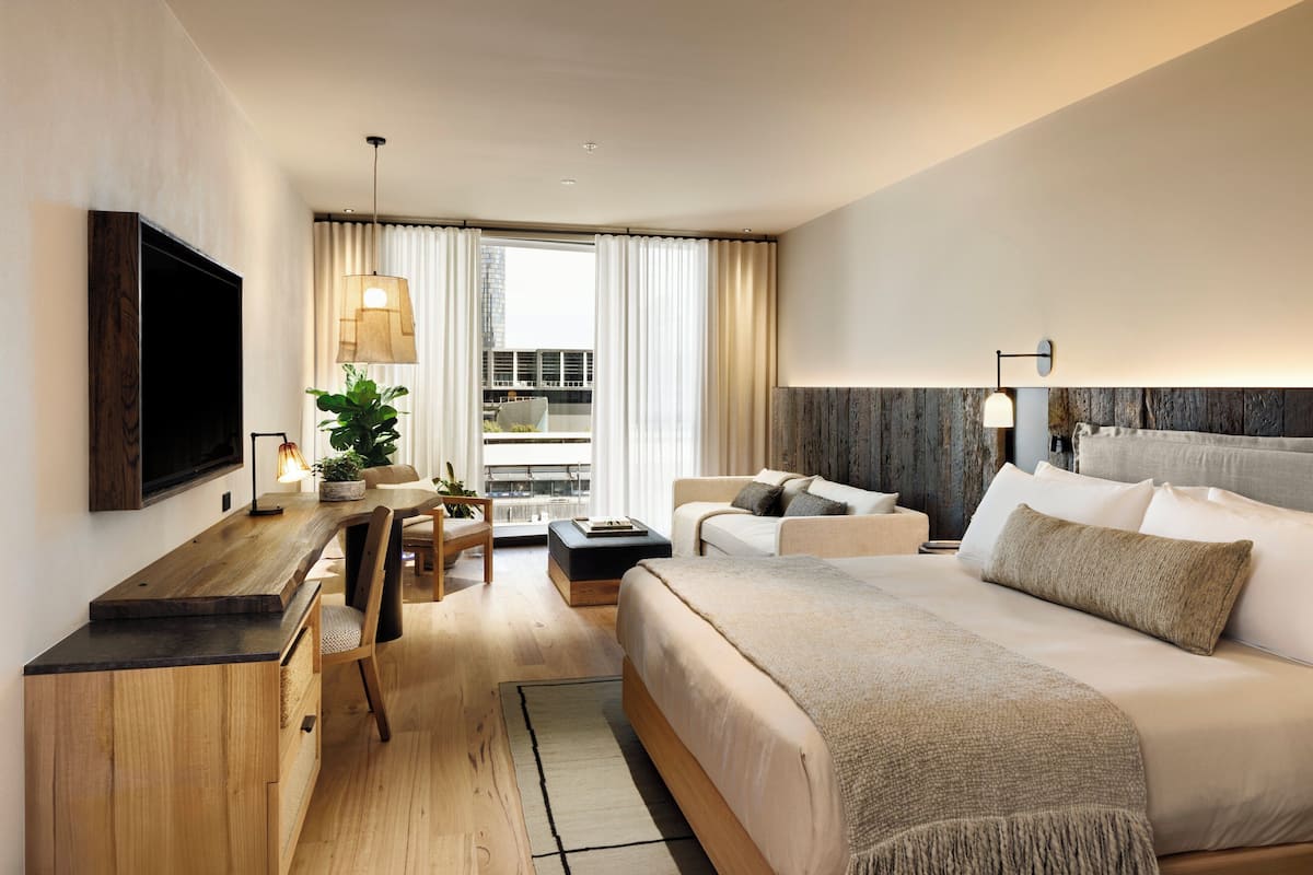 Una habitación de hotel moderna con una cama grande, un escritorio con televisión de pantalla plana, una zona de estar con sofá y sillones, y vistas de la ciudad a través de grandes ventanales.