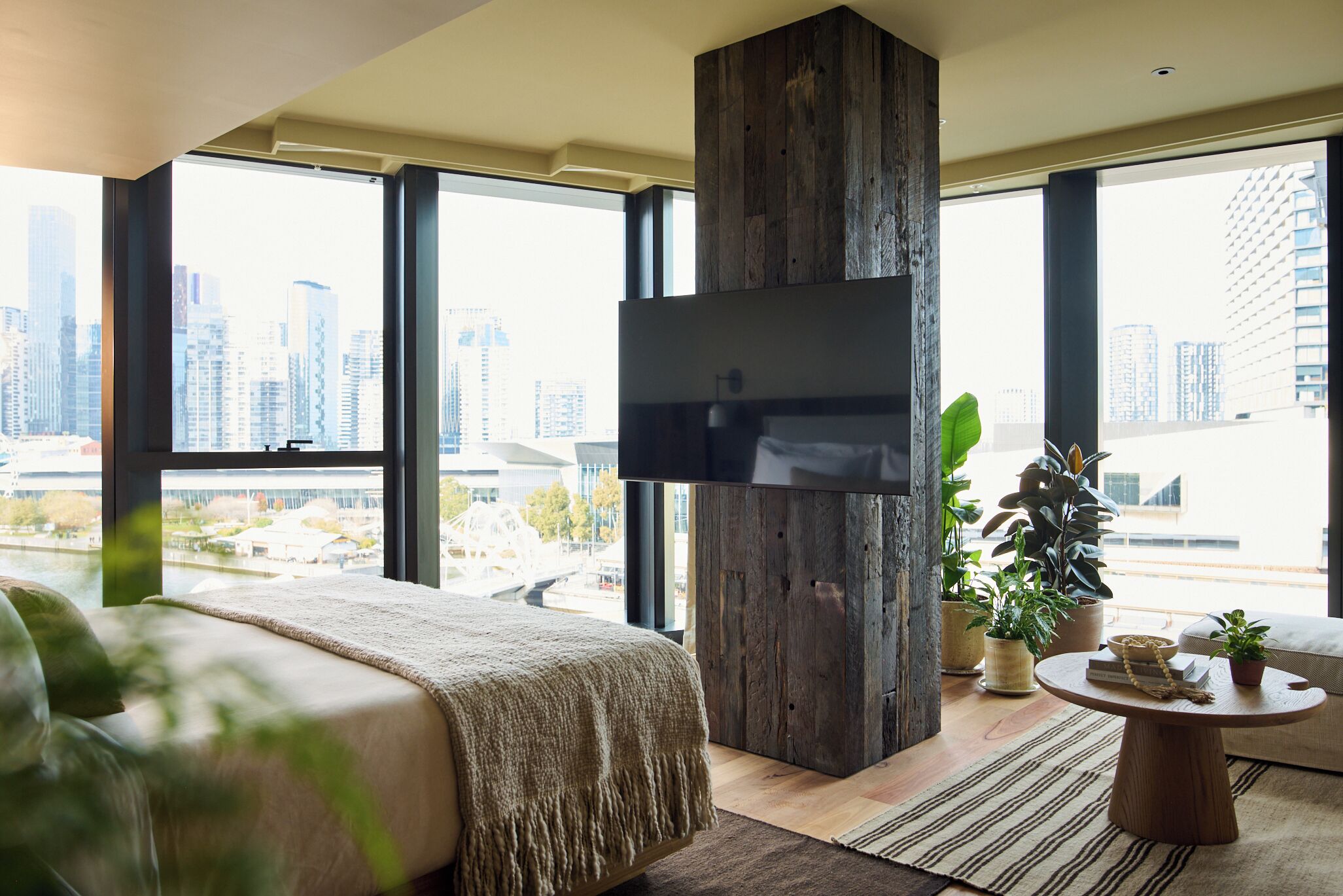 A modern bedroom with a large bed, a wooden accent wall, a TV mounted on the wall, and a view of the cityscape.
