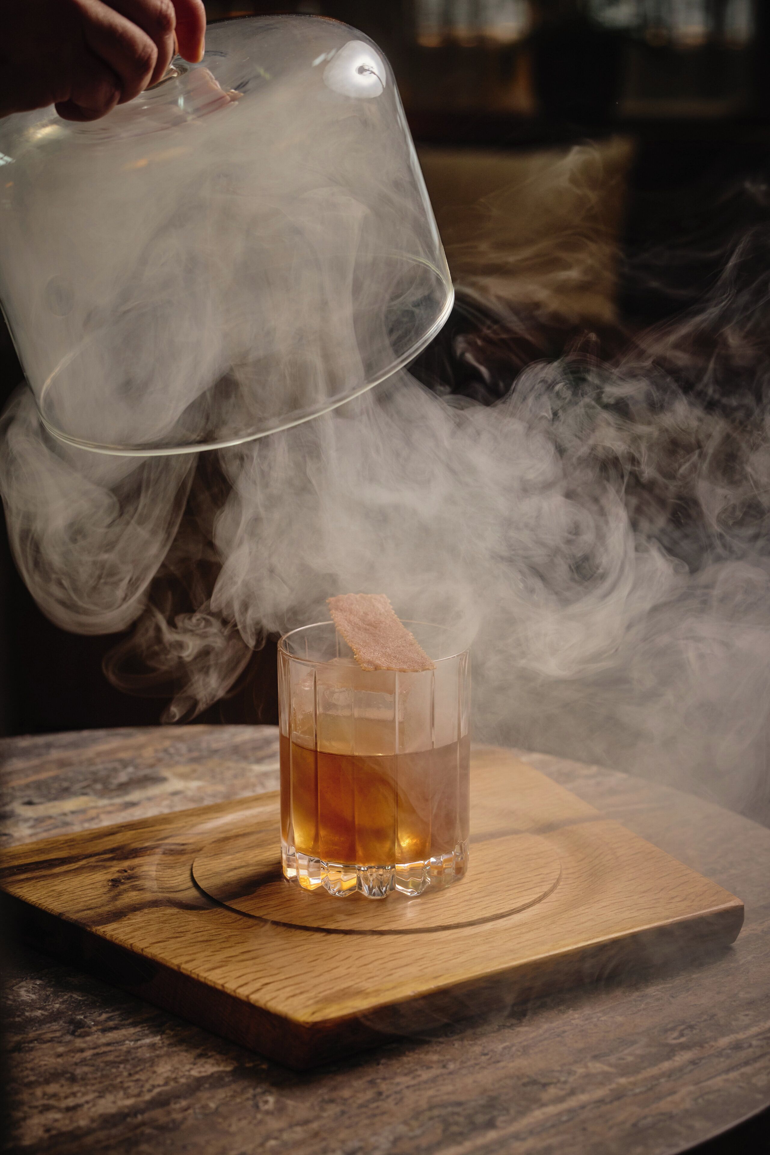 Un got amb una beguda sobre un taulell de fusta, vapor que s'eleva i una mà que sosté un recipient transparent per sobre.