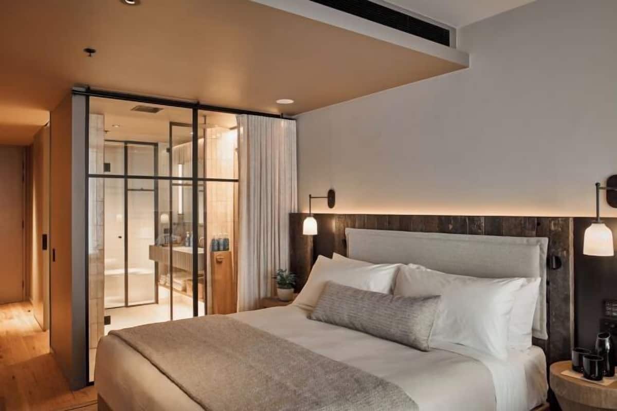 Una habitación de hotel moderna con una cama grande, un cerramiento de vidrio corredero hacia el baño y un cabecero de madera.