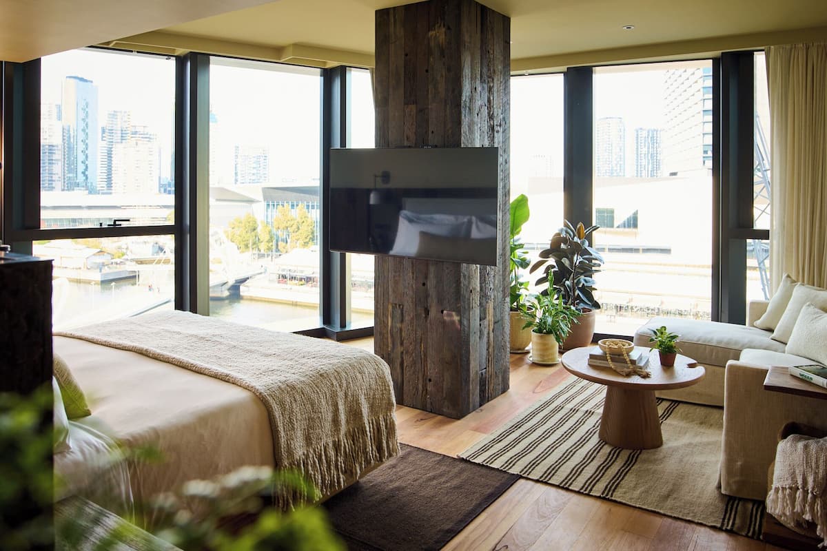 Una habitación de hotel moderna con una cama grande, un sofá, una mesa de centro y un televisor fijado a un pilar de madera.