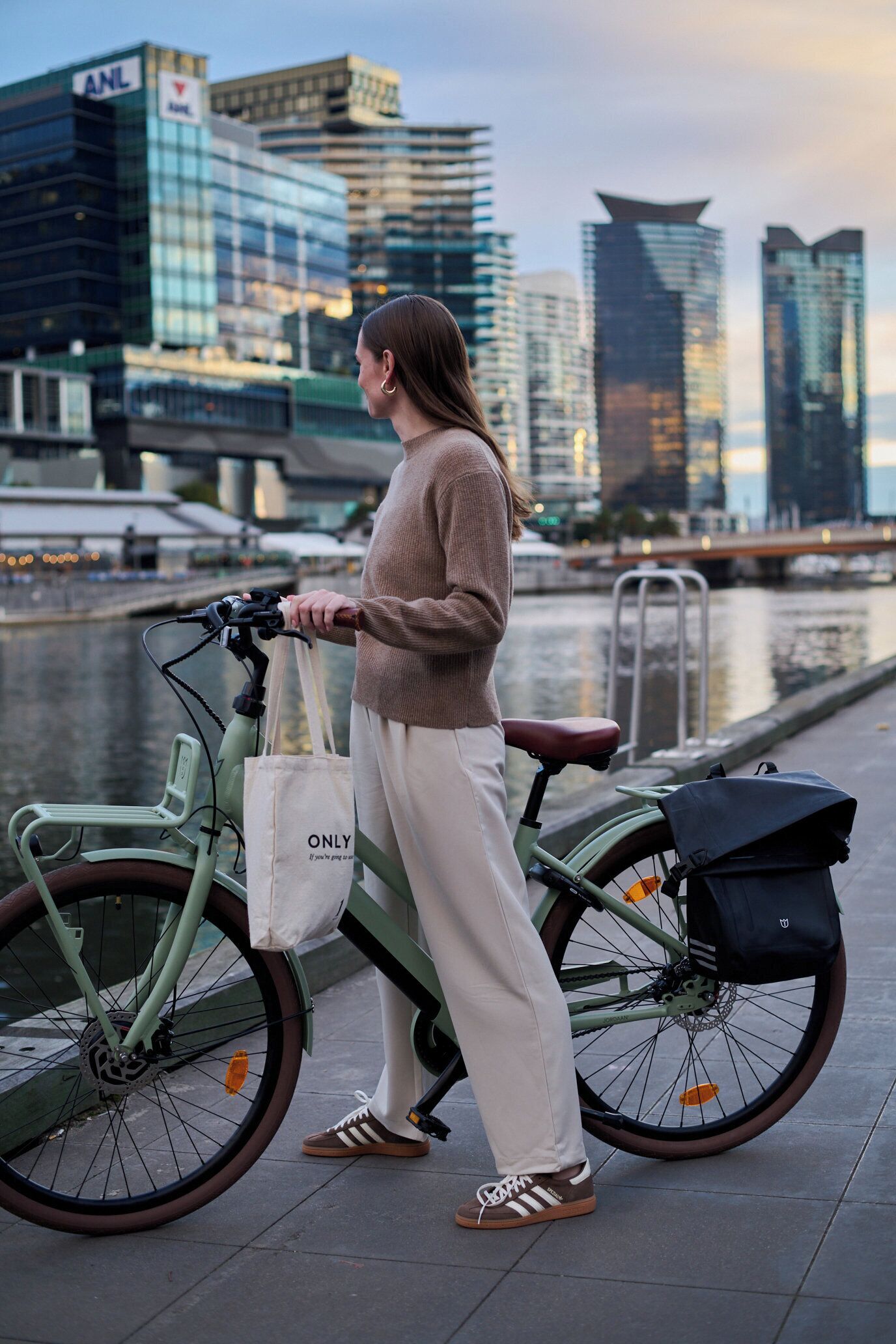 Una persona de pie junto a una bicicleta verde con un bolso blanco y uno negro, en un entorno urbano con edificios modernos y un cuerpo de agua al fondo.