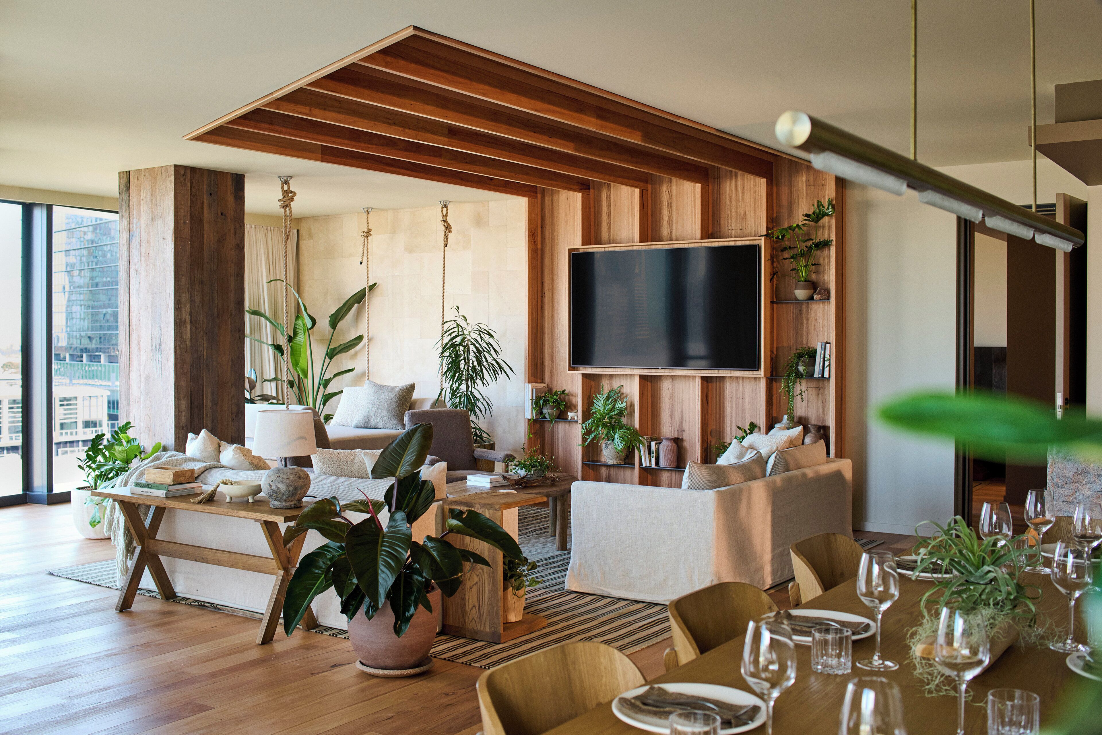 A modern living room with wooden beams, a flat-screen TV, multiple seating areas, and abundant indoor plants.