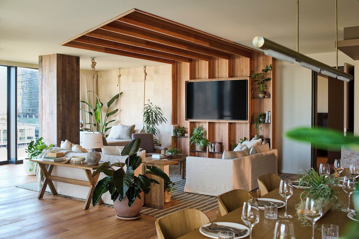 Una sala de estar moderna con techo de madera, un televisor de pantalla plana grande, varias zonas de estar y abundantes plantas de interior.