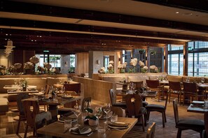 Un restaurante con muebles de madera, mesas dispuestas con copas de vino y vistas a la ciudad a través de amplios ventanales.