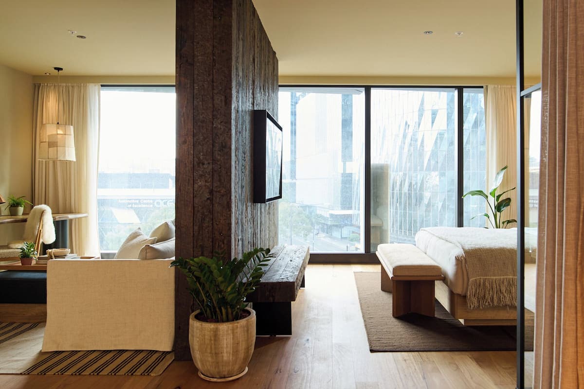 Una habitación de hotel moderna con un ventanal grande, un banco de madera, un sofá, una mesita de noche y una planta en maceta.