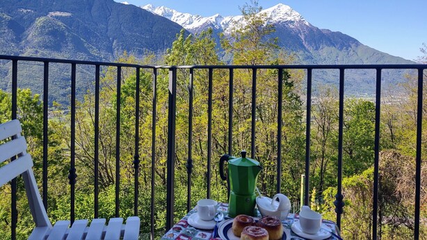 A balcony with a table set for breakfast, overlooking snow-capped mountains and lush greenery. - Casa Kiwi by Interhome (Cercino)
