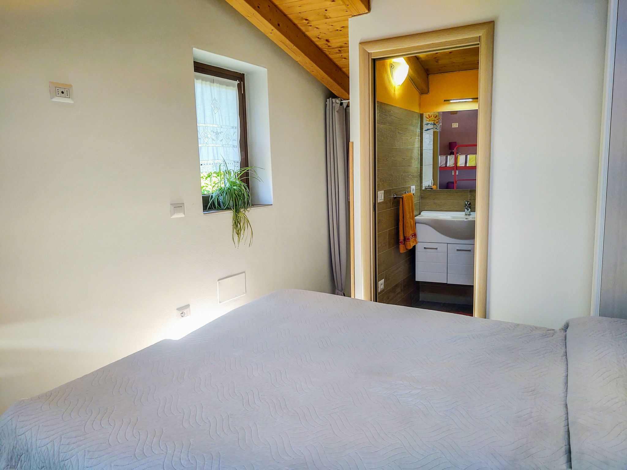 Una camera da letto con un letto, una finestra con una pianta e un bagno visibile attraverso una porta aperta.