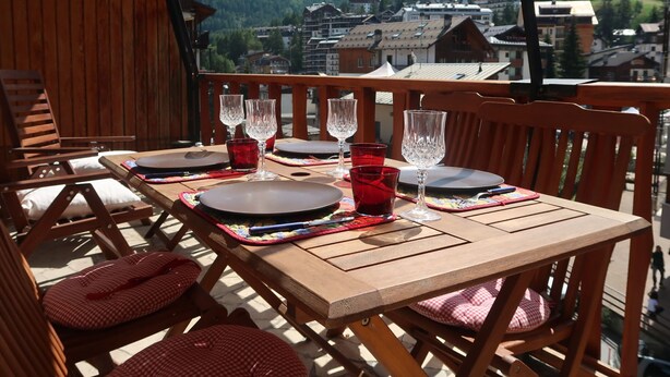 Mesa de madera preparada para cuatro personas, con vasos y platos rojos, con vistas a un pueblo montañoso.