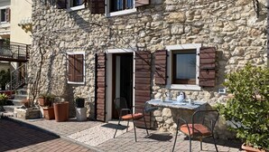 Un bâtiment en pierre avec des volets en bois, un petit espace extérieur aménagé avec une table et des chaises, et des plantes en pot.