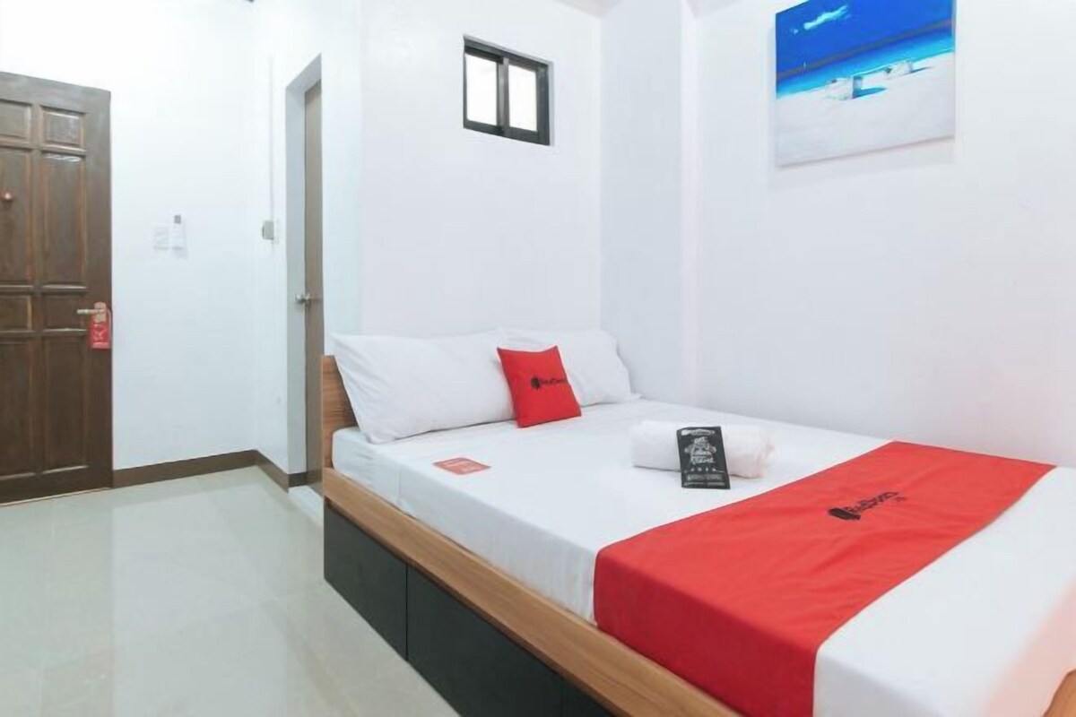 Une chambre avec un lit simple, une tête de lit en bois, du linge de lit blanc, un oreiller rouge, une petite fenêtre et un tableau accroché au mur.