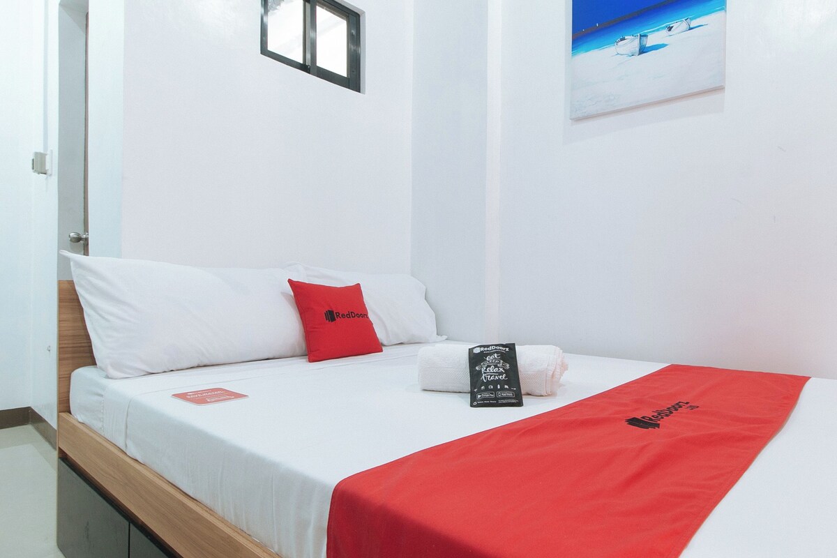 A hotel room with a bed, white bedding, a red throw, a pillow with a logo, a folded towel, a small window, and a painting on the wall.