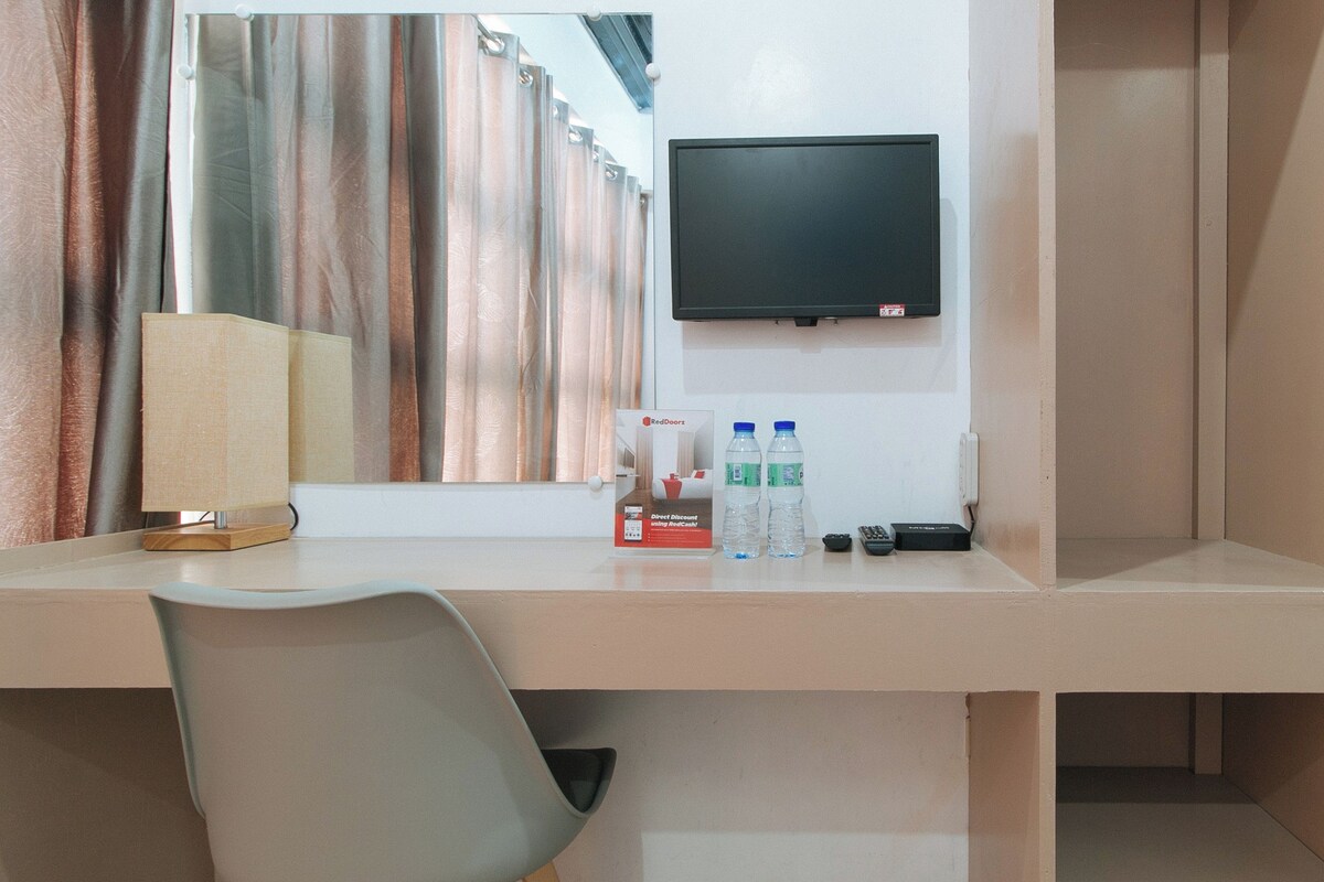 A hotel room with a desk, a television mounted on the wall, a lamp, two water bottles, and a remote control.