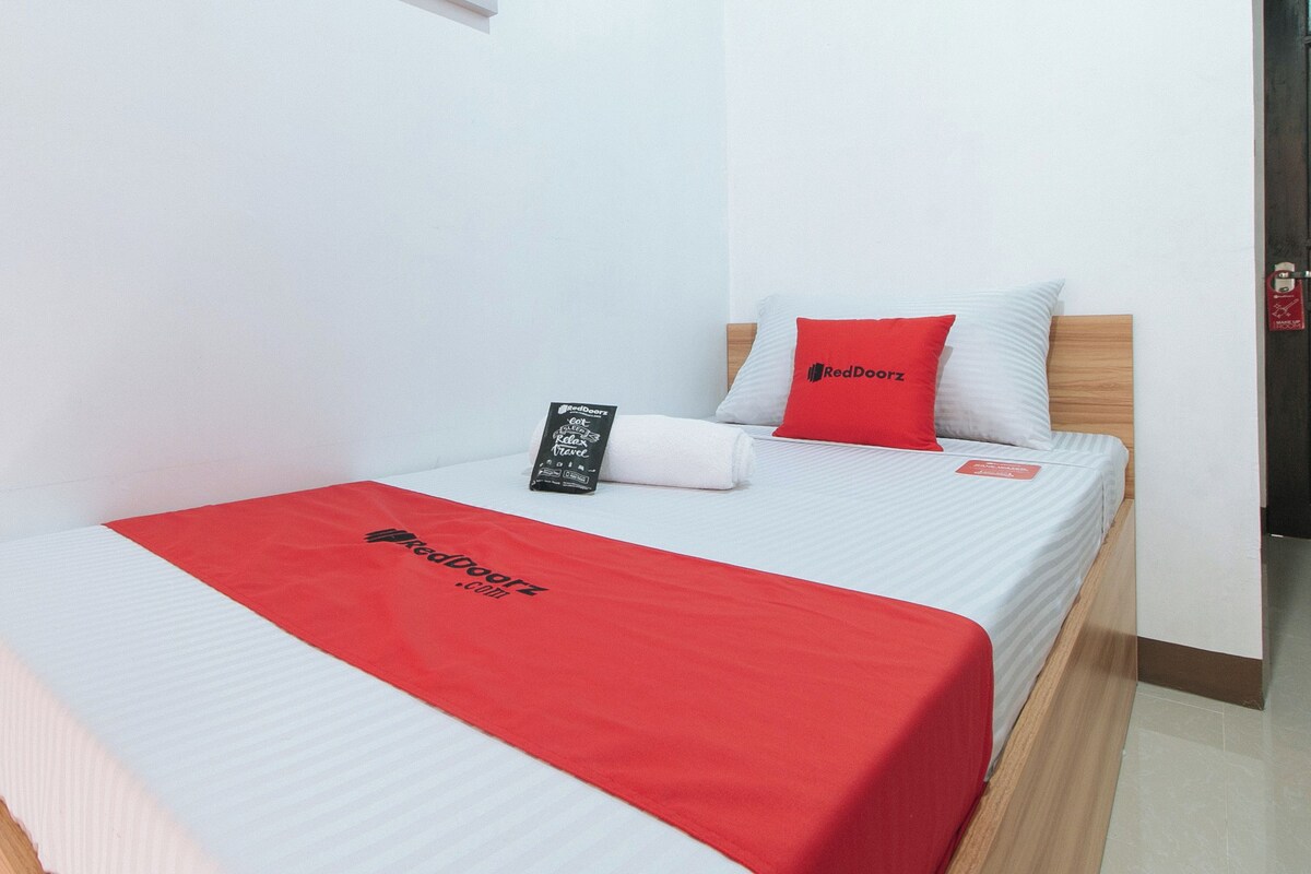 A single bed with a white and red bedspread, a red pillow, a black pillow, and a red blanket with the RedDoorz logo.