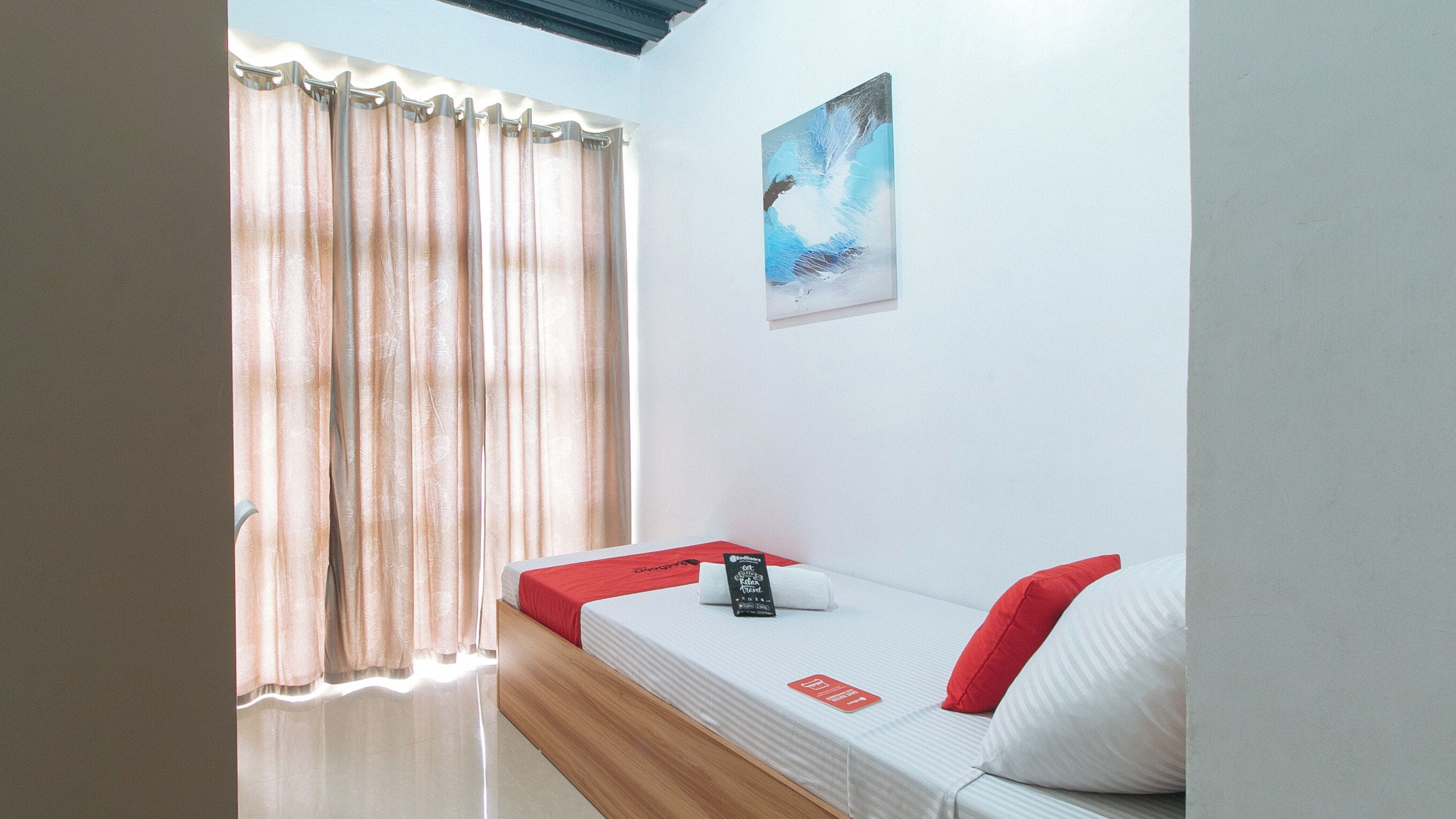 Una habitación con una cama, una ventana con cortinas, un cuadro en la pared y una almohada roja sobre la cama.