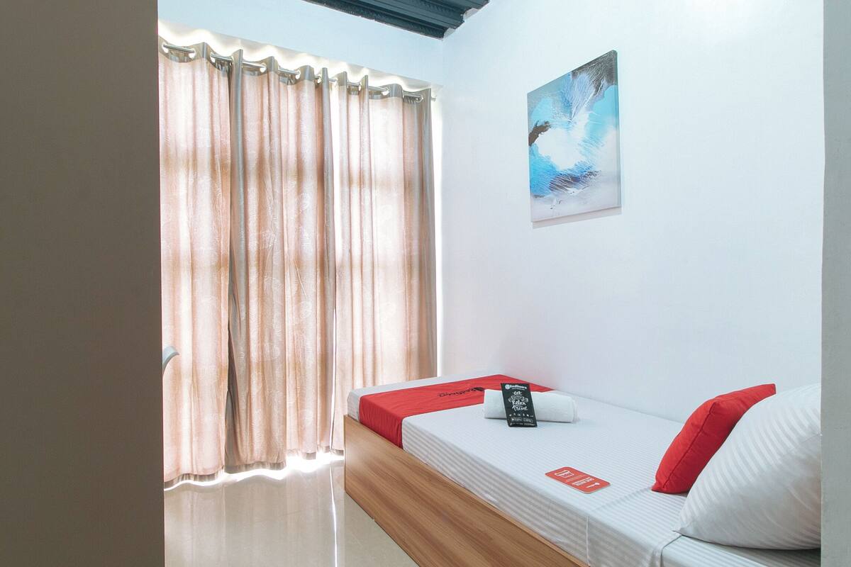 A room with a bed, a window with curtains, a painting on the wall, and a red pillow on the bed.