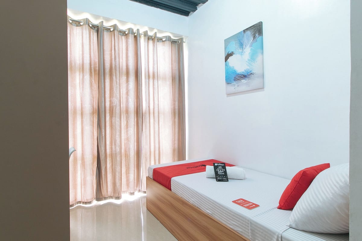 Una habitación con una cama, una ventana con cortinas, un cuadro en la pared y una almohada roja sobre la cama.