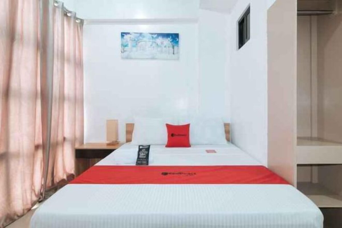A single bed with a red headboard, white bedding, and a red accent pillow. There is a small painting on the wall above the bed.