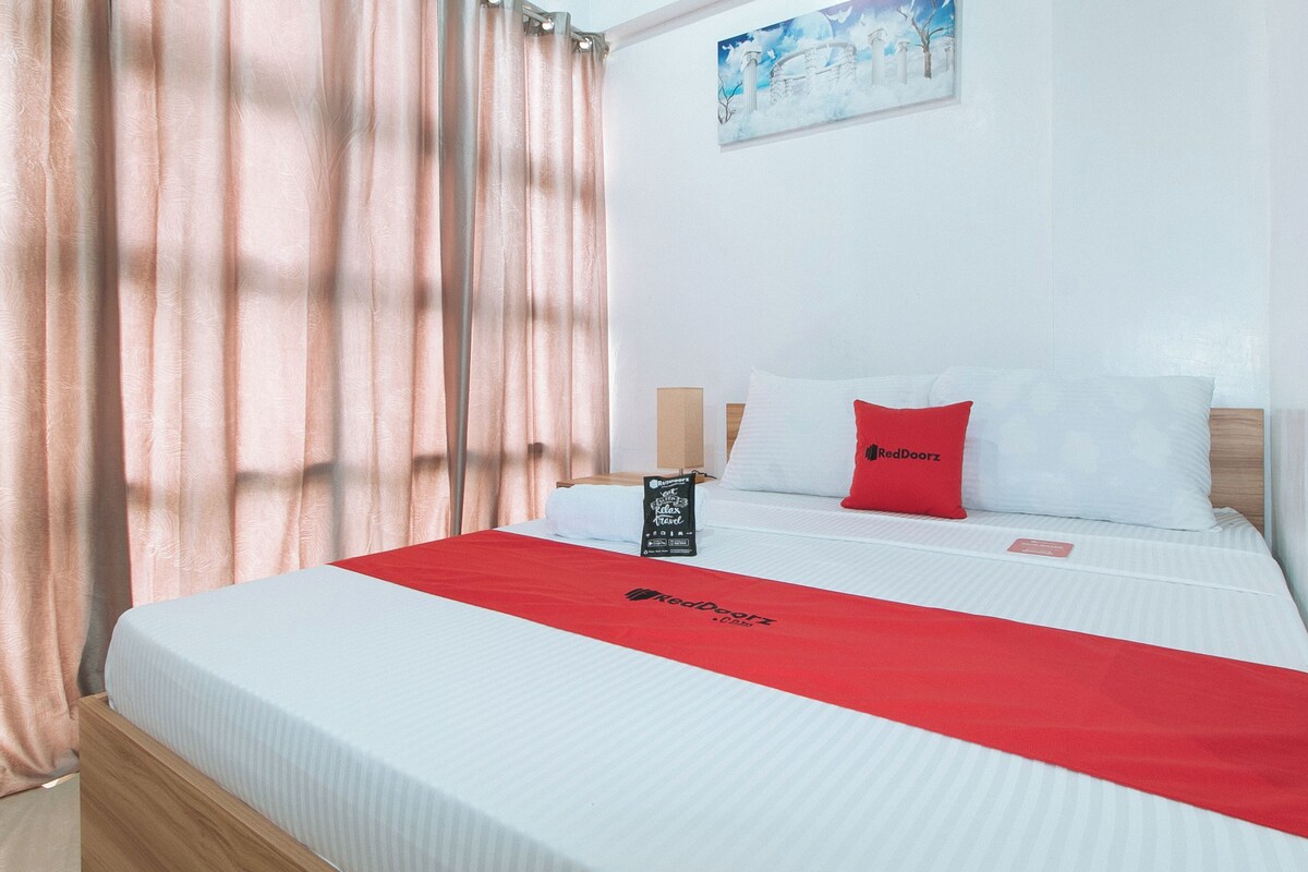 A hotel room with a bed, a red bedspread, a pillow with a logo, a nightstand with a lamp, and a painting on the wall.