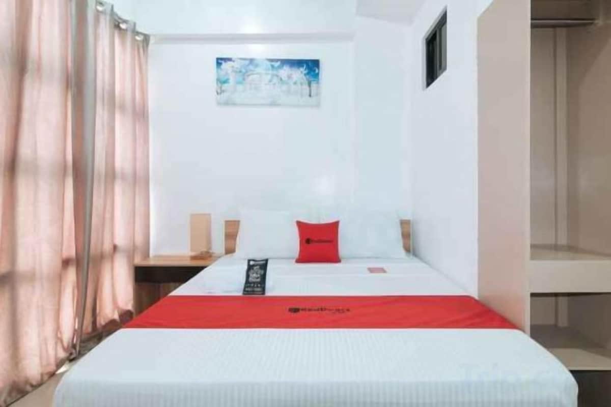 A hotel room with a bed, a red pillow, a white bedspread, a painting on the wall, and a window with curtains.