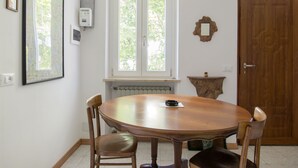 Un coin repas comprenant une table ronde en bois, quatre chaises, une fenêtre donnant sur un espace verdoyant, un radiateur placé sous la fenêtre, une prise électrique murale et une porte en bois.