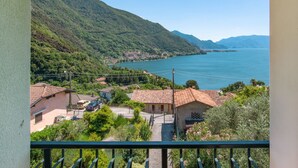 Un balcon donnant sur un lac, entouré de maisons aux toits recouverts de tuiles rouges, et un paysage montagneux en arrière-plan.