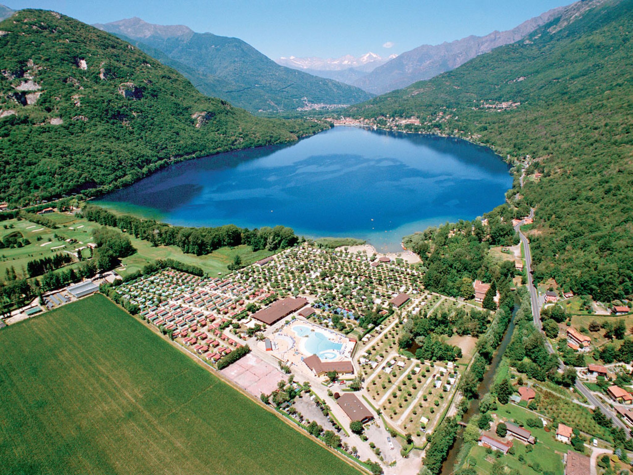 An aerial view of a resort with a large lake, surrounded by mountains, featuring a central pool area, multiple buildings, and well-maintained gardens.